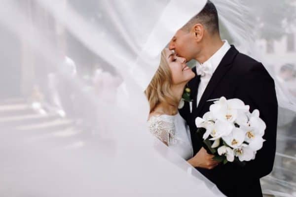 groom in black tuxedo hugs tender stunning bride while hey stand